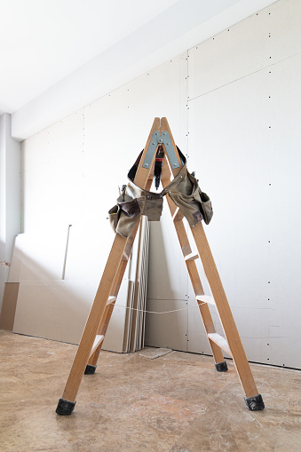 Stepladder with a tool belt hanging on it at a house construction site with plaster walls in the background. Home renovations