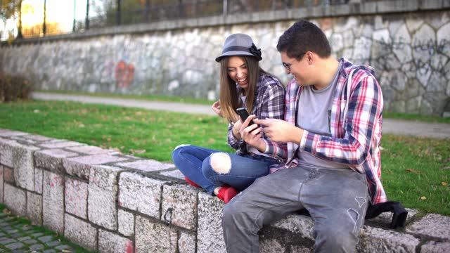 Young Couple Using Phone And Laughing While Sitting On The Quayside