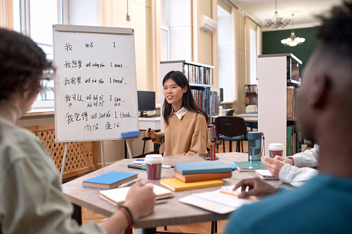 Portrait of young Asian woman as female teacher talking to group of students sitting at table by whiteboard with hieroglyphs copy space