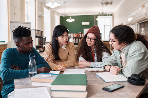 Multiethnic group of students discussing project sitting at table in college library with young Asian woman speaking