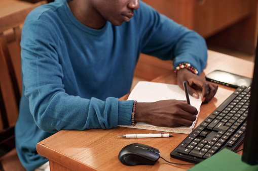 Close up of Black male student writing in notebook while using computer in college library and doing homework copy space