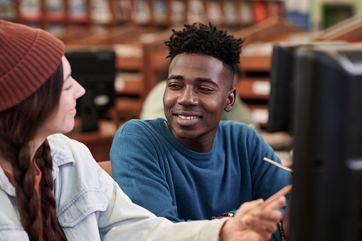 Portrait of smiling young Black man talking to friend while using computer together for studying in college library