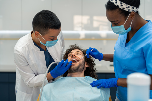 Female dentist is drilling man's tooth and healing caries while the nurse is assisting her.