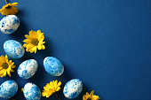 Happy Easter greeting card with painted eggs and yellow flowers on dark blue background. Top view, flat lay, copy space.
