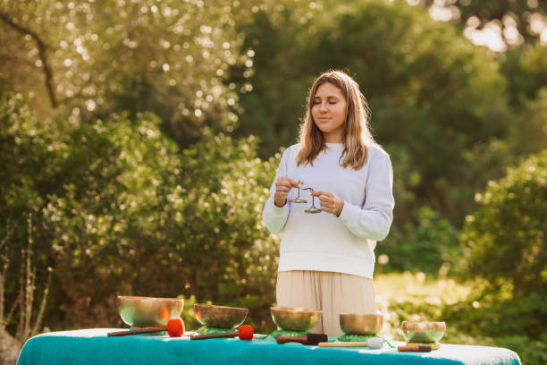 jovem mulher pacífica em pé com címbalos na frente de cinco tigelas de canto tibetano velho em seu jardim - spirituality hand on heart meditating women - fotografias e filmes do acervo