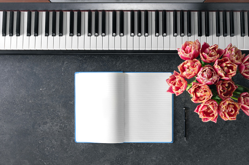 Piano, bouquet of tulip flowers and notepad on a dark background, top view. Music learning concept, copy space.