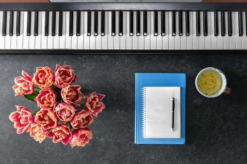 Piano, bouquet of spring flowers, notepads and a cup of tea on a dark background, top view.