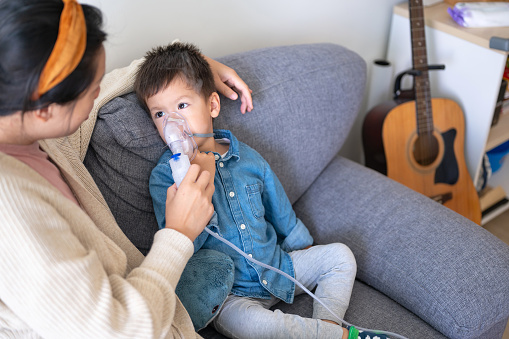 A young child uses a nebulizer for respiratory therapy while his mother assists, sat on a home couch. A child receives medical care at home with the assistance of their mother, showcasing the importance of family support in healthcare