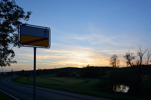 Colorful road sign on the evening highway in perspective on the background of a beautiful sky with the setting sun and fields
