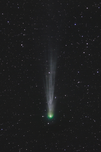 Astrophoto of periodic comet 12P/Pons-Brooks in the night sky. Green nucleus and tail of ion, dust and vapor illuminated by the sun in front of star field in space.