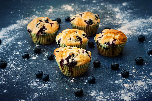 Five freshly baked blueberry muffins on a blue countertop, surrounded by blueberries and flour scattered around.