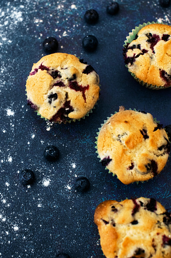 Four freshly baked blueberry muffins on a blue countertop, surrounded by blueberries and flour scattered around.
