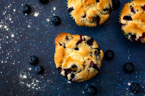 Three freshly baked blueberry muffins on a blue countertop, surrounded by blueberries and flour scattered around.
