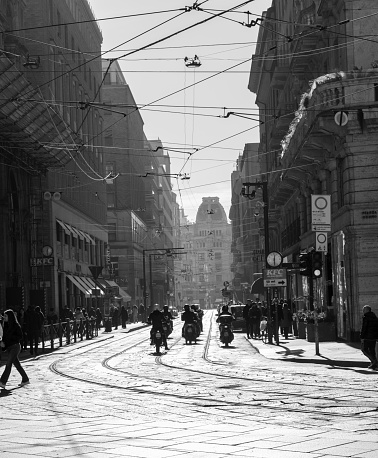 This is an image of a busy Street in the city of Milan. It was taken in the morning and shows the tramlines in the cobbles. There a lots of pedestrians and scooters.