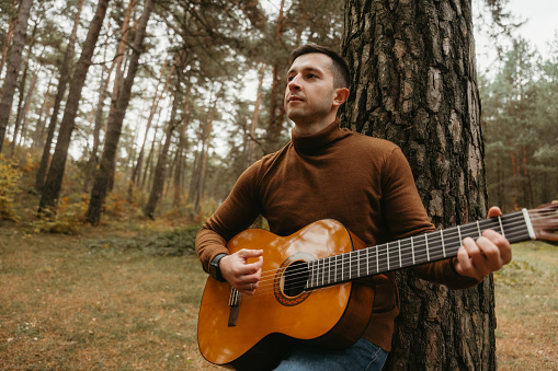Caucasian man plays guitar leaning on tree in woods, surrounded by nature