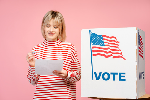 Beautiful, cheerful young woman, supporter holding documents choosing, American presidential election standing isolated on pink background. Concept vote