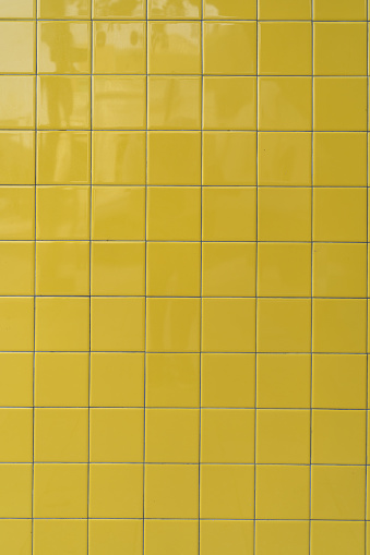 Section of wall covered with neatly arranged yellow tiles. Square-shaped material have uniform in size and color creating consistent and orderly look. Glossy surface. High quality photo