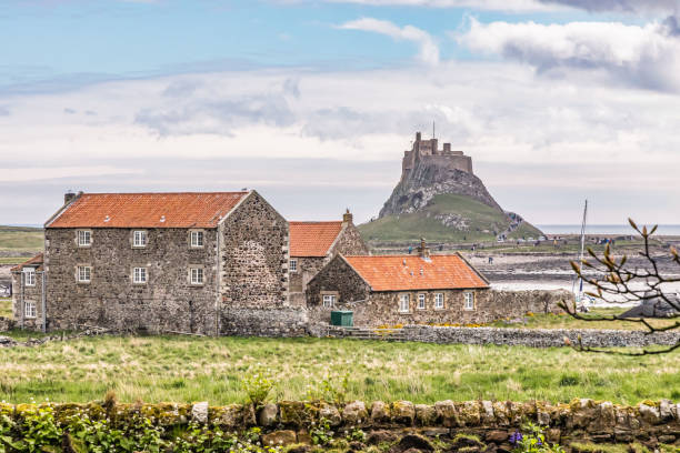 A view of Lindisfarne Castle on the Holy Isle in Northumberland, England against a pale summer sky with headland and boats in the foreground.