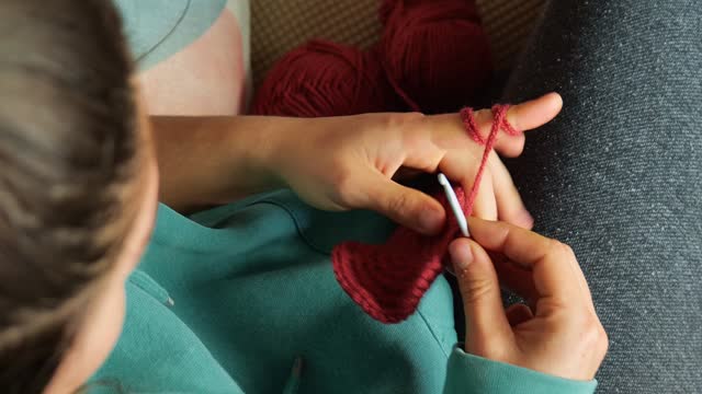 Young Caucasian woman knitting a scarf with red yarn. Sitting on the sofa in the living room, close-up over the shoulder.