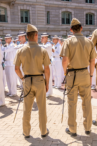 Several Naval Academy upperclassman, distinguishable by their tan uniforms and swords, monitor a formation of the incoming Plebe (freshman) midshipmen during Plebe Summer. \n\nPlebe Summer is a rigorous orientation to Naval Academy life prior to the start of the academic year.