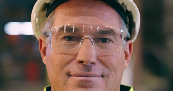 Senior man, face and safety goggles with engineer in workshop, construction or maintenance with smile.  Manager in portrait, engineering and glasses for protection in factory with labor and helmet