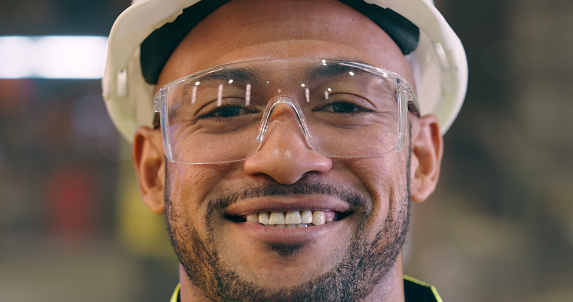 Man, face and safety goggles with engineer in workshop, construction or maintenance with smile. Employee happy in portrait, engineering and glasses for protection in factory with labor and helmet