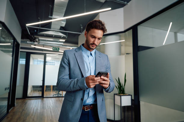 Young caucasian man in businesswear using mobile phone in corridor of office Young caucasian man wearing businesswear using mobile phone in corridor of corporate office frosted glass door office stock pictures, royalty-free photos & images