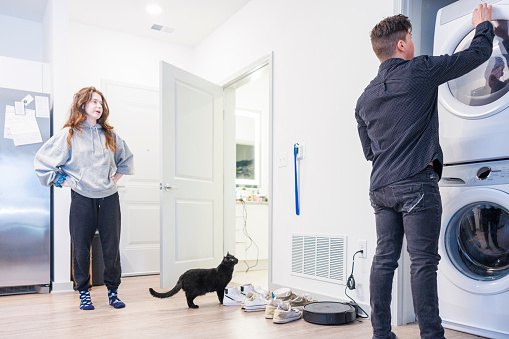 Multiracial couple of Latino man and Caucasian woman split domestic chores. Young man does the laundry and drying, while woman and cat watching over him