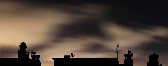 Silhouette of an urban rooftop with chimneys and television aerials at night with motion blurred clouds illuminated by city street lamps.
