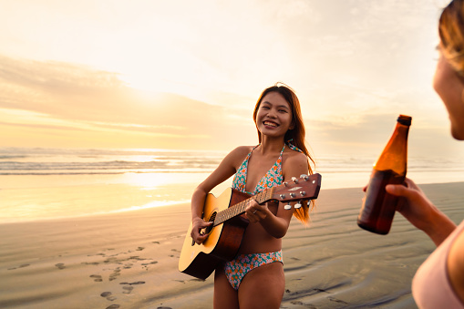 Two friends at beach enjoying and having fun one playing guitar and other having a drink, weekend activities concept .