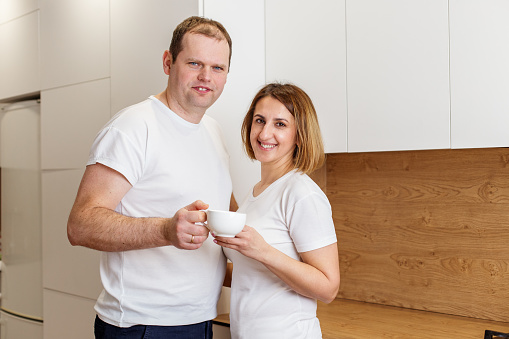 Cheerful couple stands together in their kitchen, each holding white cup of tea, enjoying relaxed morning at home.