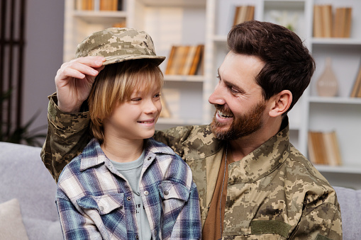 Smiling son wearing his father's army hat, sits on his dad's knees. Father, dressed in Ukrainian military gear, delighted to be back home, aims to nurture a patriotic spirit in his child