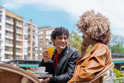 Two young friends meet for breakfast at a coffee shop, laughing and eating, Social and cultural inclusion, Hispanic, Afro Latino, in buildings and city streets in background.