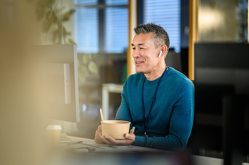 Smiling mid adult asian man in earbuds chatting in web conference on computer in office