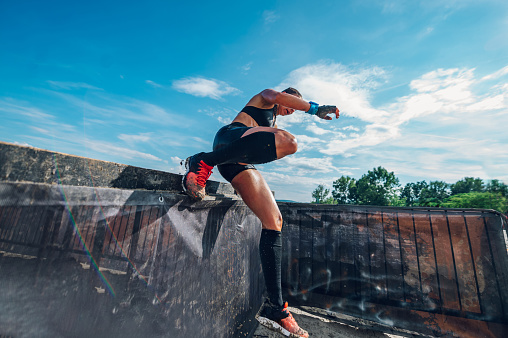 Fit sportswoman participating in an physically demanding obstacle course race and overcoming one of the obstacles. OCR race concept. Focus on a woman legs while jumping over the fence.