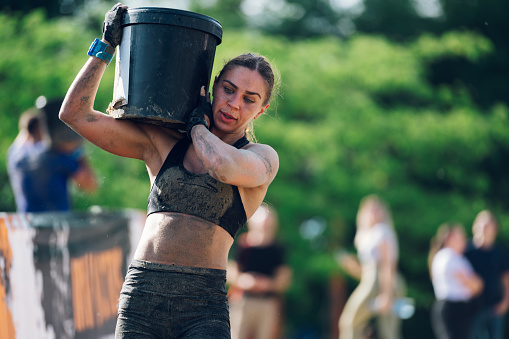 Portrait of a strong sportswoman participating in an physically demanding obstacle course race and carrying a heavy barrel resting on her shoulder. OCR race concept. Copy space.