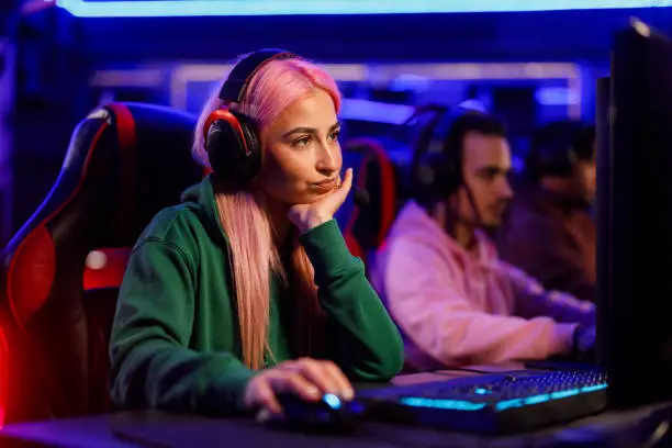 Bored woman with pink hair professional esport gamer wearing headphones and playing online video games while sitting in gaming club, internet cafe or cybersport gaming lounge. Waiting to play.