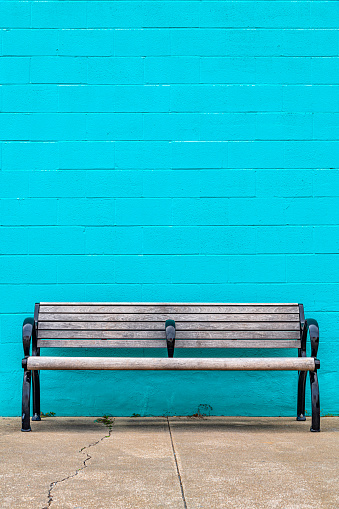 Abstract view of a municipal park bench bolted to the sidewalk shot against a blue painted concrete cinder-block wall.