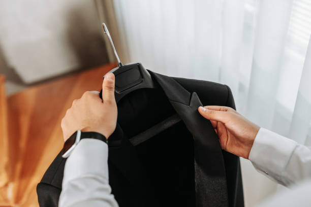 A man's hands hold a black jacket on a hanger by the window in his room.