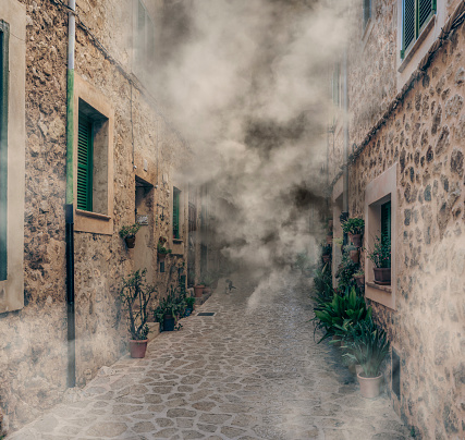 Eerie mist floating through a quaint stone street lined with potted plants.