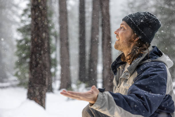 A man wearing a black hat and a blue jacket is standing in the snow, holding his hands out to catch snowflakes. Concept of joy and playfulness as the man embraces the winter weather