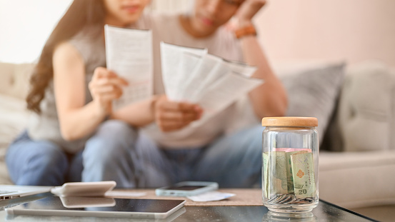 A close-up image of a jar of coins on a coffee table with a blurred background of a stressed couple worrying about their household bills and planning their finances. savings, budget, income