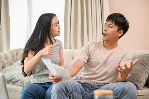 A resentful, angry Asian wife and husband are arguing about family household invoices and high domestic bills, quarreling about the issue of wasting money on the sofa in the living room.