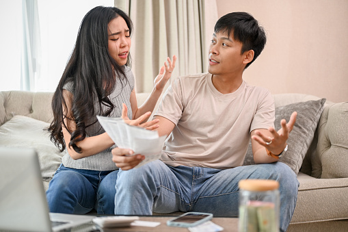 A resentful, angry Asian wife and husband are arguing about family household invoices and high domestic bills, quarreling about the issue of wasting money on the sofa in the living room.