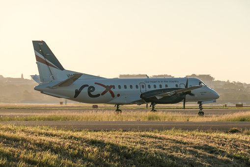 A Rex Airlines Saab 340B+ plane, registration VH-ZPCF, taxiing to the domestic terminal of Sydney Kingsford-Smith Airport after landing as flight ZL6833 from Dubbo. This image was taken from near General Holmes Drive, Mascot, on a hot, sunny day at sunset on 3 March 2024.