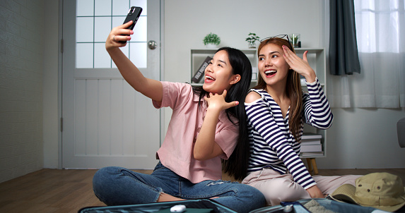 Two Asian woman use smartphone taking selfie during during packing clothes to suitcases preparing go to travel vacation, laugh with happiness together