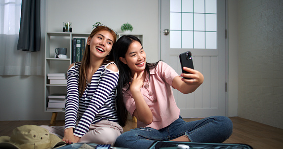 Two Asian woman use smartphone taking selfie during during packing clothes to suitcases preparing go to travel vacation, laugh with happiness together