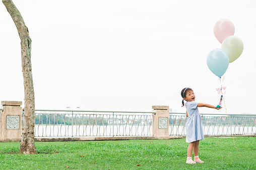 Innocence in motion! A delightful scene unfolds at the public park as a charming 3-year-old Asian baby girl giggles with joy while playing with a colorful flying balloon. Her tiny fingers reach for the sky, capturing the pure delight of childhood in this heartwarming moment of outdoor play