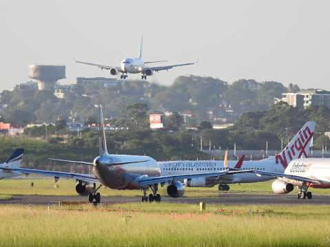 A Virgin Australia Boeing B737-8FE plane, registration VH-YIW, landing from the north at Sydney Kingsford-Smith Airport as flight VA869 from Melbourne. Four planes are visible as waiting to depart: a Jetstar A321-231, VH-VWW; a Virgin B737-7K2, VH-NVW; a Jetstar A320-232, VH-YXT; and a Rex Saab 340B.  In the left background is a concrete water tower. This image was taken from near General Holmes Drive, Mascot, on a hot and sunny evening before sunset on 3 March 2024.