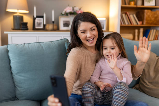 istock Smiling mother and daughter on video call using digital phone at home. while making video call and waving hello sitting on sofa. Woman and her cute little kid doing video conference with grandparents. 2062376054